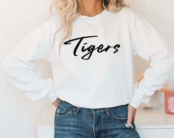 Pode incluir: Camisola branca com letras pretas em escrita cursiva que dizem "Tigers". A modelo está usando jeans azul.
