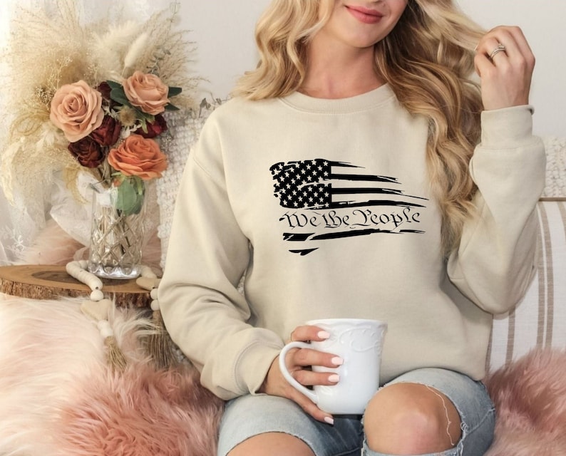 May include: A beige sweatshirt with a distressed American flag design and the text "We the People" in black.