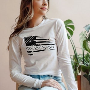 May include: White long-sleeve shirt with a distressed American flag design and the text "We the People" printed on the front.