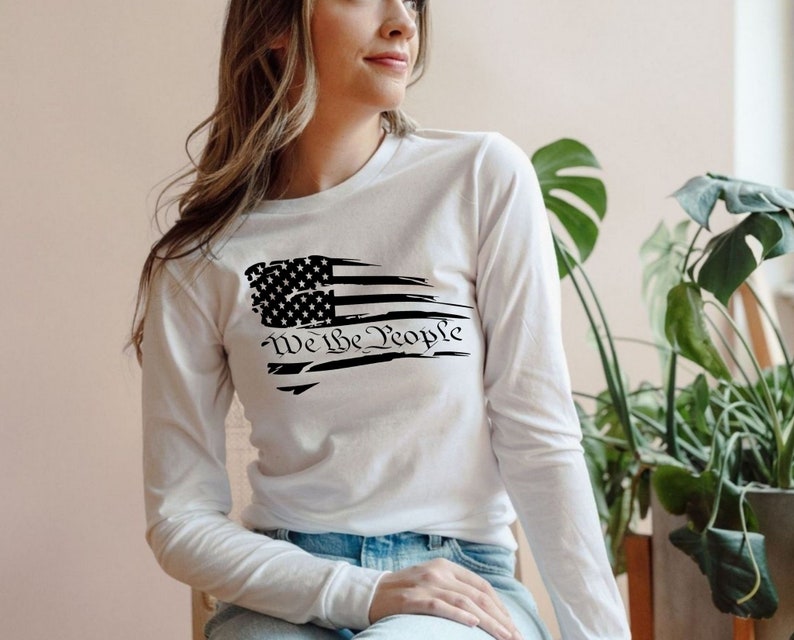 May include: White long-sleeve shirt with a distressed American flag design and the text "We the People" printed on the front.