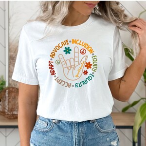 May include: White t-shirt with a colorful graphic design featuring a hand making the 'I love you' sign with the words 'Accept, Adapt, Advocate, Inclusion, Equity, Equality' surrounding the hand.