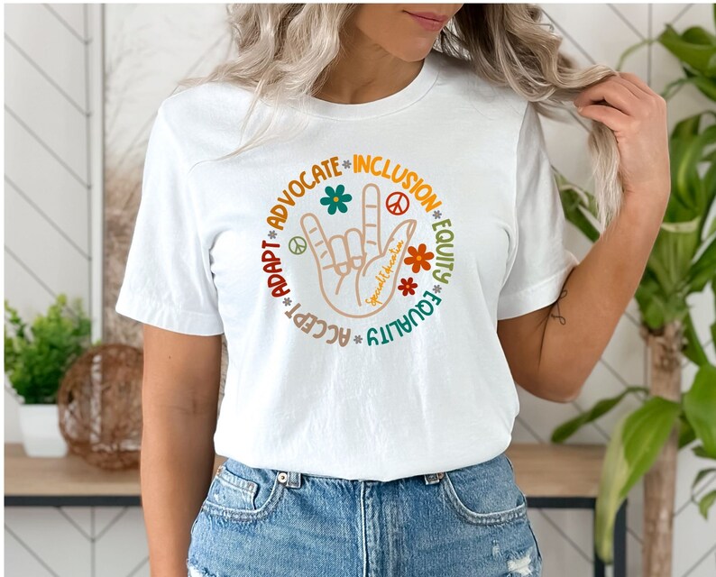May include: White t-shirt with a colorful graphic design featuring a hand making the 'I love you' sign with the words 'Accept, Adapt, Advocate, Inclusion, Equity, Equality' surrounding the hand.