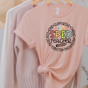 Pode incluir: Uma camiseta rosa com um design circular que diz "SPED teacher" em letras coloridas. O c&iacute;rculo &eacute; cercado por palavras como "dedicado", "amig&aacute;vel", "solid&aacute;rio", "carinhoso", "motivador", "confi&aacute;vel" e "destac&aacute;vel".