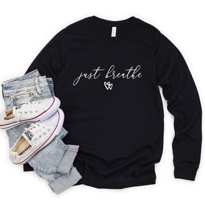 May include: Black long-sleeved t-shirt with the text "just breathe" in white script with two white heart symbols. The shirt is paired with blue jeans and white trainers.
