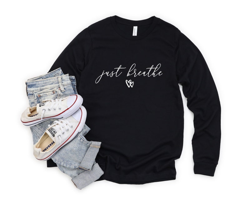 May include: Black long-sleeved t-shirt with the text "just breathe" in white script with two white heart symbols. The shirt is paired with blue jeans and white trainers.