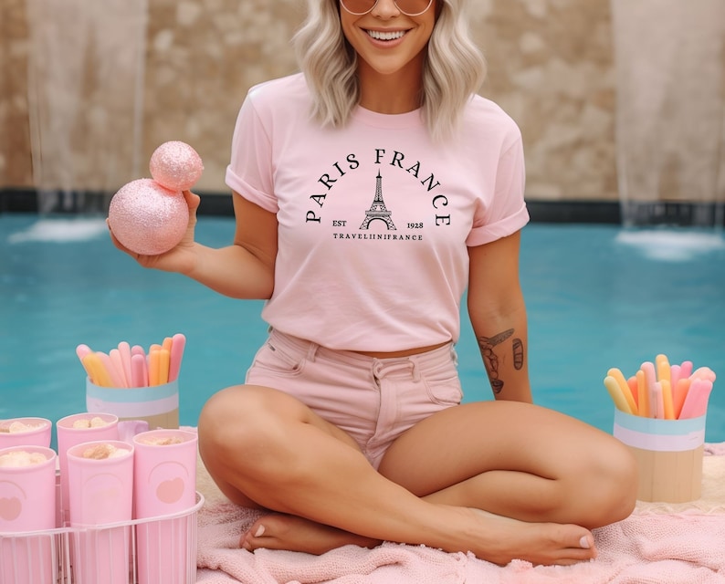 May include: A woman wearing a pink t-shirt with a black graphic of the Eiffel Tower and the text "Paris France Est 1958 TravelinInFrance". She is sitting by a pool with pink pool floats and a pink and white striped towel.