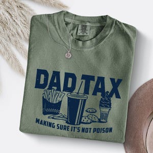 K&ouml;nnte beinhalten: Olivgr&uuml;nes T-Shirt mit dem marineblauen Schriftzug "DAD TAX" und Illustrationen von Pommes, einem Getr&auml;nk, Keksen und Eis. Der Text "MAKING SURE IT'S NOT POISON" steht darunter. Eine silberne Halskette liegt auf dem Shirt.