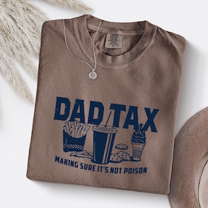 K&ouml;nnte beinhalten: Ein braunes T-Shirt mit dem navyblauen Schriftzug "DAD TAX" und Illustrationen von Pommes, einem Getr&auml;nk, Keksen und Eis. Der Text "MAKING SURE IT'S NOT POISON" steht darunter. Eine silberne Halskette liegt auf dem Shirt.