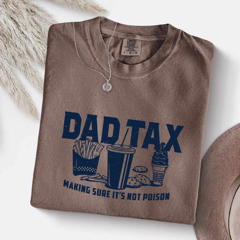 K&ouml;nnte beinhalten: Ein braunes T-Shirt mit dem navyblauen Schriftzug "DAD TAX" und Illustrationen von Pommes, einem Getr&auml;nk, Keksen und Eis. Der Text "MAKING SURE IT'S NOT POISON" steht darunter. Eine silberne Halskette liegt auf dem Shirt.