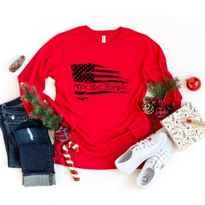 May include: Red long-sleeve shirt with a distressed American flag graphic and the text "We the People" in black.