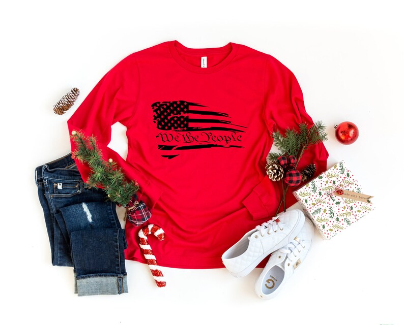 May include: Red long-sleeve shirt with a distressed American flag graphic and the text "We the People" in black.