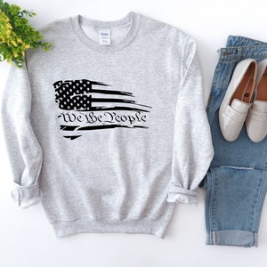 May include: A gray sweatshirt with a distressed American flag design and the text "We the People" printed in black.