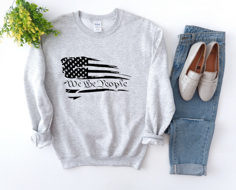 May include: A gray sweatshirt with a distressed American flag design and the text "We the People" printed in black.