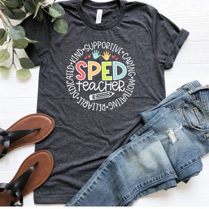 Pode incluir: Uma camiseta cinza escura com um design circular que diz "SPED teacher" em letras coloridas. O c&iacute;rculo &eacute; cercado pelas palavras "dedicado, gentil, solid&aacute;rio, atencioso, motivador, confi&aacute;vel".