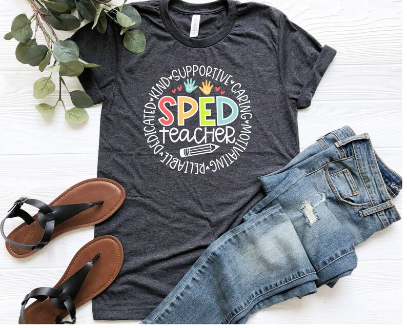 Pode incluir: Uma camiseta cinza escura com um design circular que diz "SPED teacher" em letras coloridas. O c&iacute;rculo &eacute; cercado pelas palavras "dedicado, gentil, solid&aacute;rio, atencioso, motivador, confi&aacute;vel".