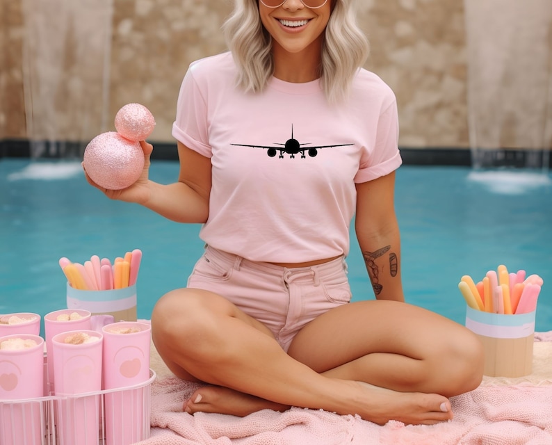 May include: A pink t-shirt featuring a black silhouette of an airplane with the word "Plane" printed on the front. The person wearing the shirt is sitting by a swimming pool.