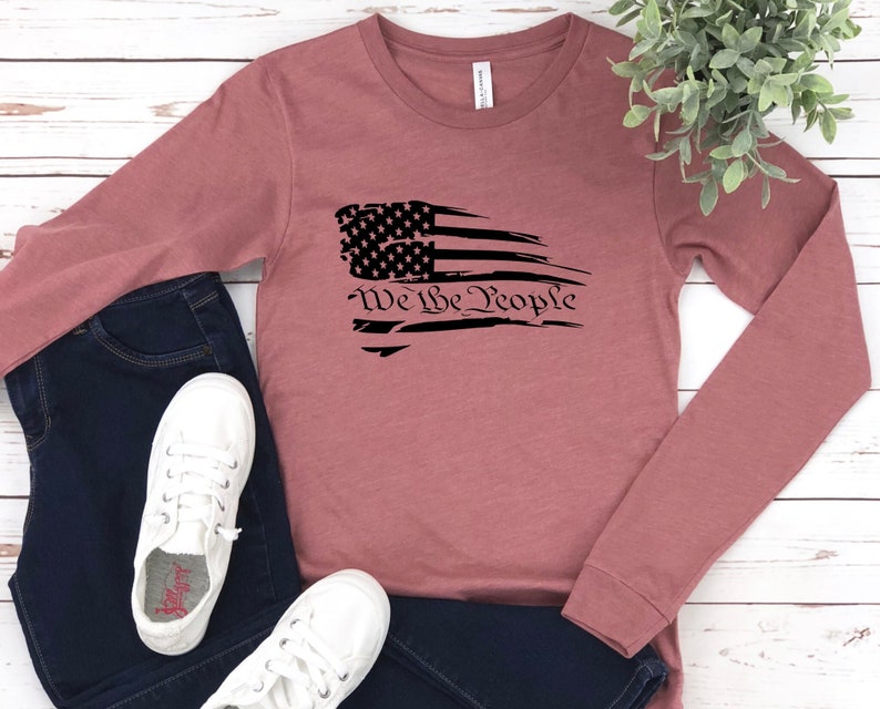 May include: A long-sleeved, mauve t-shirt with a distressed black and white American flag graphic and the text "We the People".