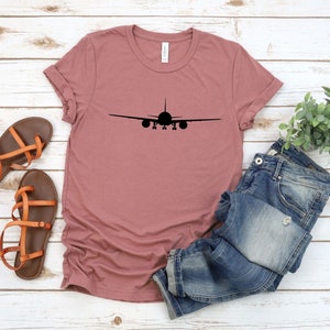 May include: A mauve t-shirt with a black airplane silhouette printed on the front. The shirt is laid out on a white wooden surface with a pair of brown sandals and blue jeans.