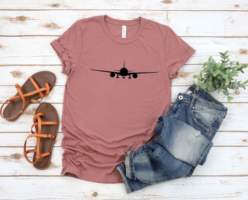 May include: A mauve t-shirt with a black airplane silhouette printed on the front. The shirt is laid out on a white wooden surface with a pair of brown sandals and blue jeans.