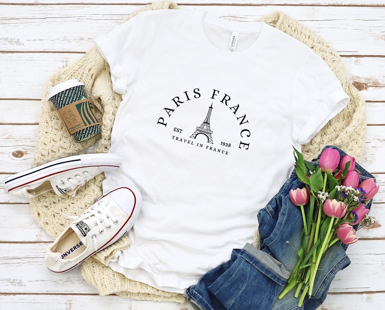 May include: White t-shirt with a black graphic of the Eiffel Tower and the text "Paris France Est 1928 Travel in France".