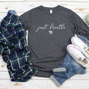 May include: A dark grey long-sleeved t-shirt with the text "just breathe" in white script with two white hearts above the text. The shirt is laid out on a white wooden surface with a plaid shirt, blue jeans, and white trainers.