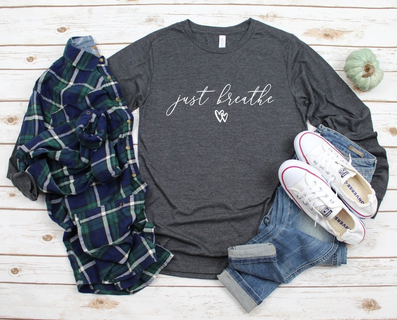 May include: A dark grey long-sleeved t-shirt with the text "just breathe" in white script with two white hearts above the text. The shirt is laid out on a white wooden surface with a plaid shirt, blue jeans, and white trainers.