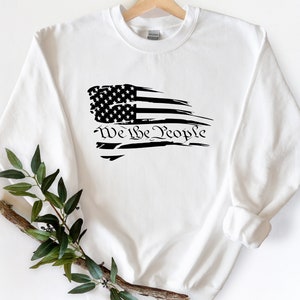 May include: White sweatshirt with a black distressed American flag design and the text "We the People" printed below the flag.