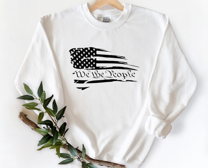 May include: White sweatshirt with a black distressed American flag design and the text "We the People" printed below the flag.