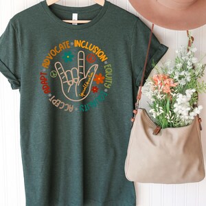May include: A dark green t-shirt with a circular design featuring the American Sign Language symbol for "I love you" surrounded by the words "Advocate", "Inclusion", "Equity", "Accept", "Adapt", "Listen", and "Spread kindness".