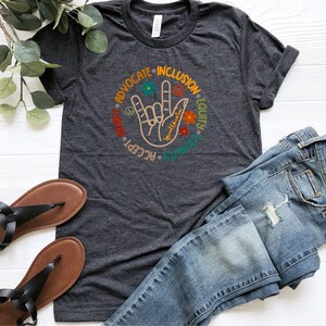 May include: A dark gray t-shirt with a graphic of a hand making the American Sign Language sign for "I love you". The graphic is surrounded by a circle of words: "Advocate", "Inclusion", "Equity", "Respect", "Accept", "Unity", "Love", "Peace", and "Kindness".