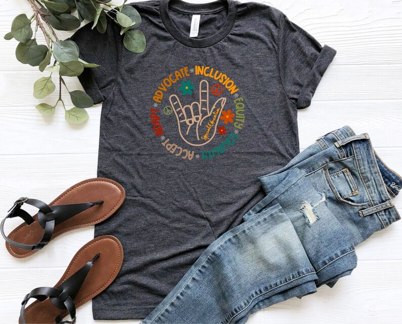 May include: A dark gray t-shirt with a graphic of a hand making the American Sign Language sign for "I love you". The graphic is surrounded by a circle of words: "Advocate", "Inclusion", "Equity", "Respect", "Accept", "Unity", "Love", "Peace", and "Kindness".