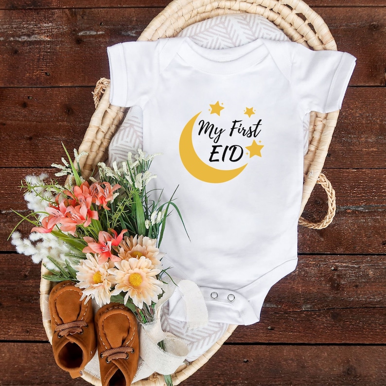 K&ouml;nnte beinhalten: Wei&szlig;er Baby-Strampler mit einem gelben Halbmond- und Sternen-Design. Der Text "My First Eid" ist auf dem Strampler aufgedruckt.