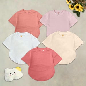 May include: Five short-sleeved t-shirts in various colors: peach, light pink, white, cream, and coral. The shirts are arranged in a circular pattern. A plush cloud with a smiling face and a sun is in the lower left corner.