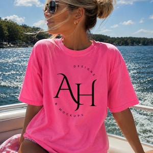 Op de afbeelding: Een felroze t-shirt met de tekst "DESIGNS & MOCKUPS" en de letters "AH" in een zwarte, gestileerde letter. Het t-shirt wordt gedragen door een persoon op een boot, met een zonnebril en een knotkapsel. De achtergrond toont een blauw meer en lucht.
