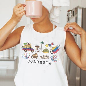 May include: White sleeveless tank top with a colorful graphic design featuring a Colombian flag, a coffee cup, a soccer ball, and the word "COLOMBIA".