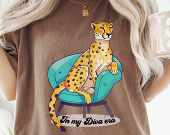 May include: A brown t-shirt with a graphic of a cheetah sitting on a teal chair. The text "In my Diva era" is printed below the cheetah.
