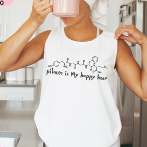 May include: White tank top with the text "pilates is My happy hour" in black. The shirt features a molecular structure design above the text. The image also includes a pink mug and a vase of pink flowers.