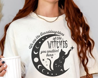May include: Ivory t-shirt with a black graphic of a crescent moon, stars, and a cat. The text says "We are the Granddaughters of the WITCHES you couldn't burn." The shirt has a loose fit. The colour is ivory.
