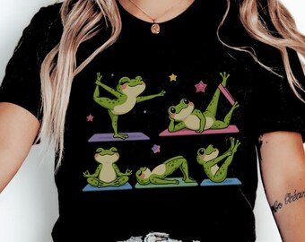 May include: A black t-shirt featuring a graphic print of five green frogs in various yoga poses on colorful mats. The frogs are depicted performing different stretches and positions, surrounded by small yellow and pink star icons.