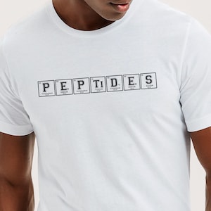 May include: A white short-sleeved t-shirt featuring a graphic print of the word PEPTIDES spelled out using individual chemical element boxes from the periodic table, each containing a letter, element name, and atomic number in black ink.