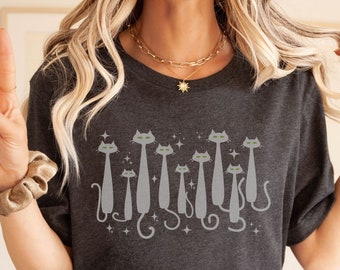 Puede incluir: Una camiseta gris oscuro jaspeada con un gráfico de una fila de gatos grises con ojos verdes. Los gatos están todos mirando hacia adelante y tienen las colas enroscadas alrededor de sus patas. El gráfico está rodeado de pequeñas estrellas blancas.