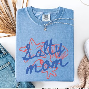 May include: A blue denim t-shirt with the text "Salty Mom" in blue and red lettering. The text is surrounded by a starfish and seashells.