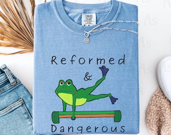 May include: A light blue t-shirt with a green frog doing a yoga pose on a bar. The text "Reformed & Dangerous" is printed on the shirt.
