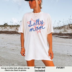 May include: White t-shirt with a blue and red graphic that says "Salty Mom" with a starfish and seashell design. The shirt is on a model wearing it. The background is a beach.