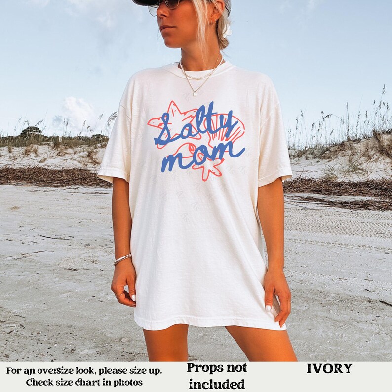 May include: White t-shirt with a blue and red graphic that says "Salty Mom" with a starfish and seashell design. The shirt is on a model wearing it. The background is a beach.