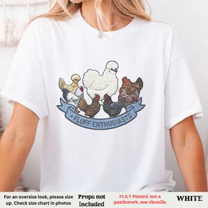 Pode incluir: T-shirt branco com uma ilustra&ccedil;&atilde;o colorida de galinhas e o texto "FLUFF ENTHUSIASTS" em uma faixa azul. Um estilo casual e confort&aacute;vel.