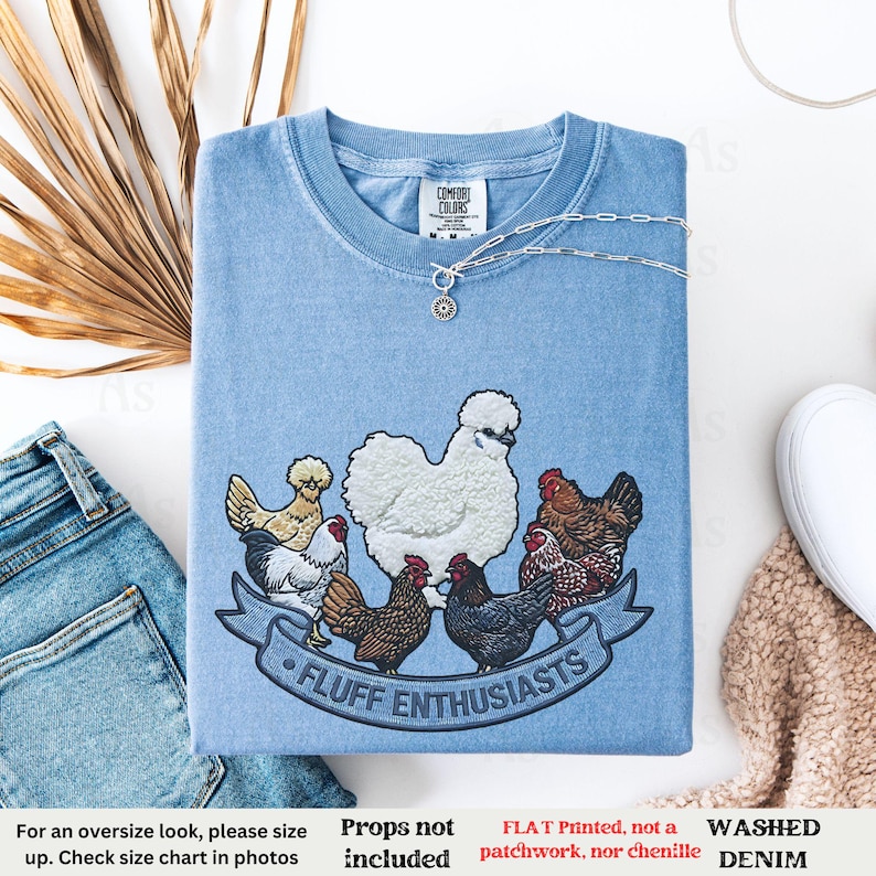 Pode incluir: Camiseta de ganga azul clara lavada com gola redonda. A camisa apresenta um design bordado de v&aacute;rias galinhas e o texto "FLUFF ENTHUSIASTS" em uma faixa. Um colar de prata com um pingente est&aacute; drapeado sobre a camisa.