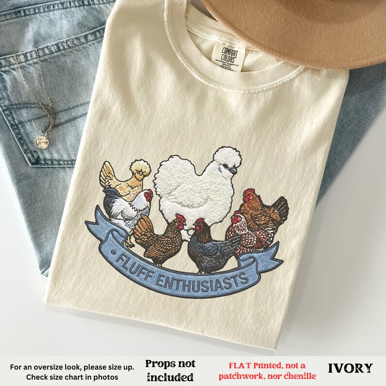Pode incluir: Camiseta marfim com um gr&aacute;fico de v&aacute;rias galinhas e o texto "FLUFF ENTHUSIASTS" em uma faixa azul. A camiseta est&aacute; dobrada sobre jeans azuis e um chap&eacute;u bege.