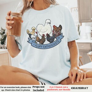 Pode incluir: Camiseta azul clara com um gr&aacute;fico colorido de v&aacute;rias galinhas e o texto "FLUFF ENTHUSIASTS" em uma faixa azul. A camiseta &eacute; feita de um material macio e &eacute; um item de roupa casual.