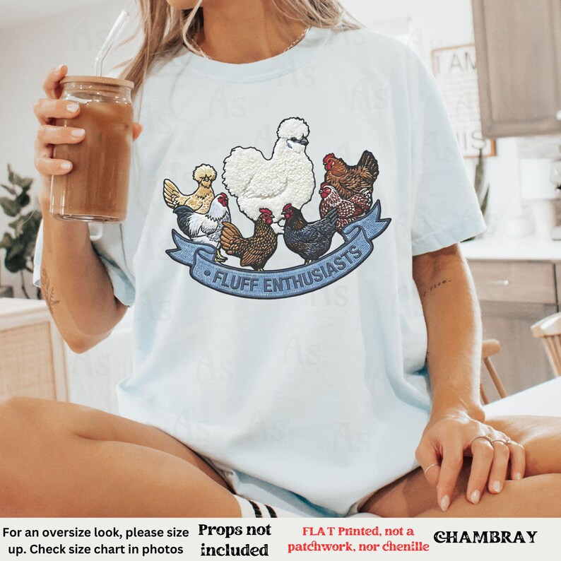 Pode incluir: Camiseta azul clara com um gr&aacute;fico colorido de v&aacute;rias galinhas e o texto "FLUFF ENTHUSIASTS" em uma faixa azul. A camiseta &eacute; feita de um material macio e &eacute; um item de roupa casual.